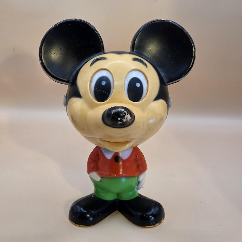 May include: Vintage Mickey Mouse figurine with black ears, a yellow face, and a red shirt. The figure has green pants and black shoes. The figurine is a classic Disney collectible.