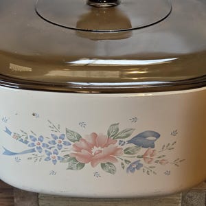 May include: A cream-colored oval casserole dish with a dark brown translucent glass lid. The exterior of the dish features a floral pattern with a central pink flower, smaller blue flowers, green leaves, and light blue ribbons on the front side.
