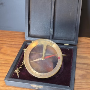 May include: A vintage brass compass with a red needle, housed in a black leatherette case with a burgundy velvet lining. The compass has a clear glass cover and a small key attached by a chain. The case is open, revealing the compass.