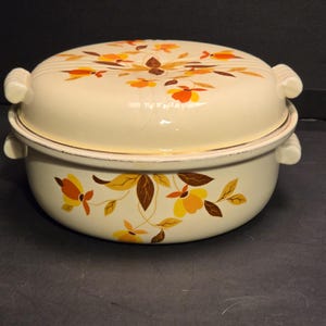 May include: A cream-colored enameled metal casserole dish with a matching lid. The exterior features a floral pattern in shades of orange, yellow, and brown. The dish has two side handles, and the lid also includes two matching side handles.