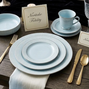 May include: Elegant table setting featuring light blue Noritake Fidelity china with silver trim. Includes plates, a teacup and saucer, a bowl, and gold-colored flatware. A place card with the name "Noritake Fidelity" is also present.