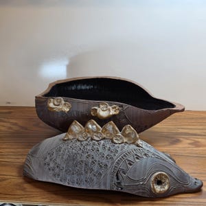 May include: A handcrafted ceramic decorative bowl with a lid. The bowl is brown with gold accents and a dark interior. The lid is gray with intricate patterns and gold embellishments. The bowl is designed to resemble a fish.