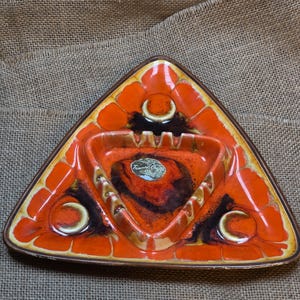 May include: A triangular ceramic ashtray featuring a vibrant orange and dark brown glaze with a textured, crackled finish. The piece includes three circular indentations and a central basin with a sticker labeled Treasure Craft.