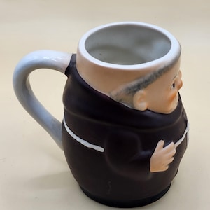 May include: A ceramic mug shaped like a monk, featuring a brown robe with a white trim and a light blue handle. The mug has a detailed face and hands, with a beige interior.