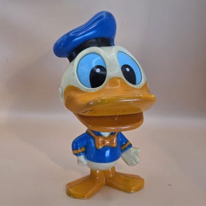 May include: A vintage Donald Duck figurine. The figure is primarily orange, white, and blue, with a blue sailor hat and a blue and white sailor suit. The figurine has large black eyes and an orange beak. The figure is a collectible item.