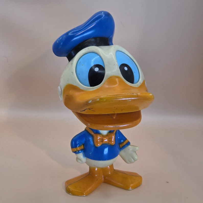 May include: A vintage Donald Duck figurine. The figure is primarily orange, white, and blue, with a blue sailor hat and a blue and white sailor suit. The figurine has large black eyes and an orange beak. The figure is a collectible item.