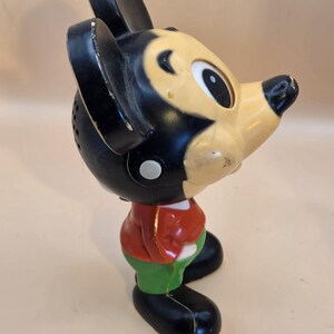 May include: Vintage Mickey Mouse figurine in a standing pose. The figure has a black head and ears, a cream-colored face, a red jacket, green pants, and black shoes. The toy is made of plastic and shows signs of wear.