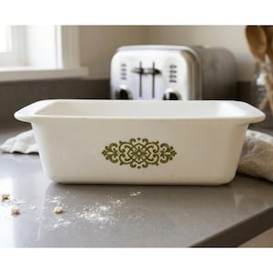 May include: A rectangular off-white ceramic loaf pan featuring a centered olive green decorative filigree pattern. The baking dish has integrated handles on both ends and a smooth, speckled finish, designed for oven use in a kitchen setting.
