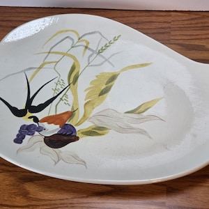 May include: An off-white, asymmetrical ceramic serving platter featuring a painted illustration of a black swallow bird, green wheat stalks, and a cluster of purple and orange fruit. The dish has a smooth, glossy finish and irregular edges.