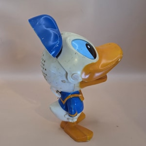 May include: Vintage Donald Duck figurine. The figure features a white head with blue accents, a yellow beak and feet, and a blue sailor suit. The figurine is made of plastic and shows signs of wear.