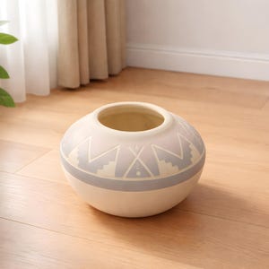 May include: A cream-colored ceramic vase with a round body and a wide opening. The vase features a gray band around the middle and a geometric pattern in gray and white. The vase sits on a wooden floor.