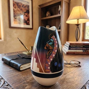 May include: A decorative ceramic vase with a Native American design. The vase features a black, white, and cream base, with a detailed illustration of a Native American figure in vibrant colors. The vase is on a wooden table.
