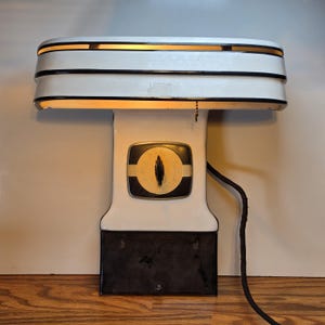 May include: Vintage white and black desk lamp with a rectangular shade and a decorative dial on the front. The lamp has a black power cord and is sitting on a wooden surface. The lamp is lit, emitting a warm glow.