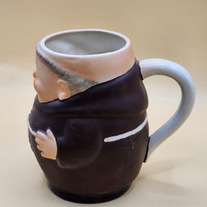 May include: A ceramic mug shaped like a monk, featuring a brown robe with white trim and a light gray handle. The mug has a tan face with a bald head and a small hand. The interior of the mug is white.