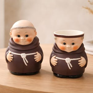 May include: Two ceramic monk figurines. One is a solid figure, the other is a planter. Both are brown with white rope belts, rosy cheeks, and smiling faces. The planter has a white interior.