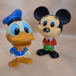 May include: Two vintage Disney character figurines: Donald Duck and Mickey Mouse. Donald Duck is in a blue sailor suit and hat, with an orange beak and feet. Mickey Mouse is in red shorts and a red shirt with black ears.