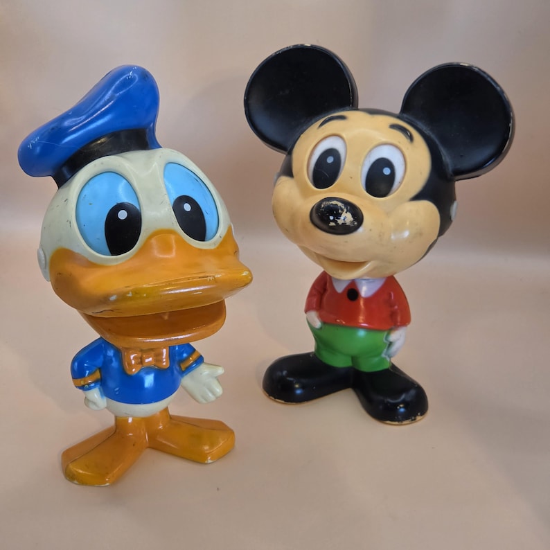 May include: Two vintage Disney character figurines: Donald Duck and Mickey Mouse. Donald Duck is in a blue sailor suit and hat, with an orange beak and feet. Mickey Mouse is in red shorts and a red shirt with black ears.