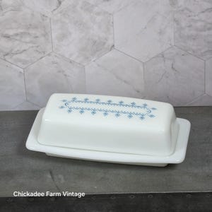 May include: A white rectangular butter dish with a lid featuring a blue decorative pattern of small star-like shapes and scalloped lines. The base has a matching white finish. The text Chickadee Farm Vintage is visible at the bottom.