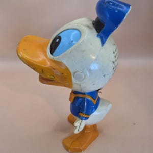 May include: Vintage Donald Duck figurine. The figure is primarily white, with a yellow beak and feet, and a blue sailor suit and hat. The figurine has black eyes and is slightly worn, showing signs of age.
