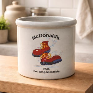 Puede incluir: Un tarro de cerámica color crema con el logotipo de McDonald's y un gráfico de botas rojas. El tarro tiene una abertura ancha y una superficie ligeramente texturizada. El texto en el tarro dice "2008 Red Wing, Minnesota."