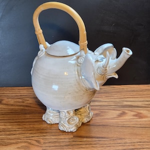 May include: A ceramic teapot in the shape of an elephant, with a light beige glaze. The teapot has a bamboo handle and is resting on four elephant-shaped legs. The trunk forms the spout. Decorative floral details are on the legs and sides.