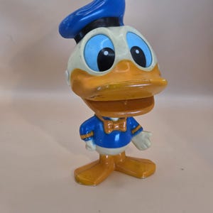 May include: A vintage Donald Duck figurine. The figure has a blue sailor hat, a white face, and a large orange beak. He wears a blue sailor shirt with a bow tie and white gloves. The figurine is standing on orange feet.