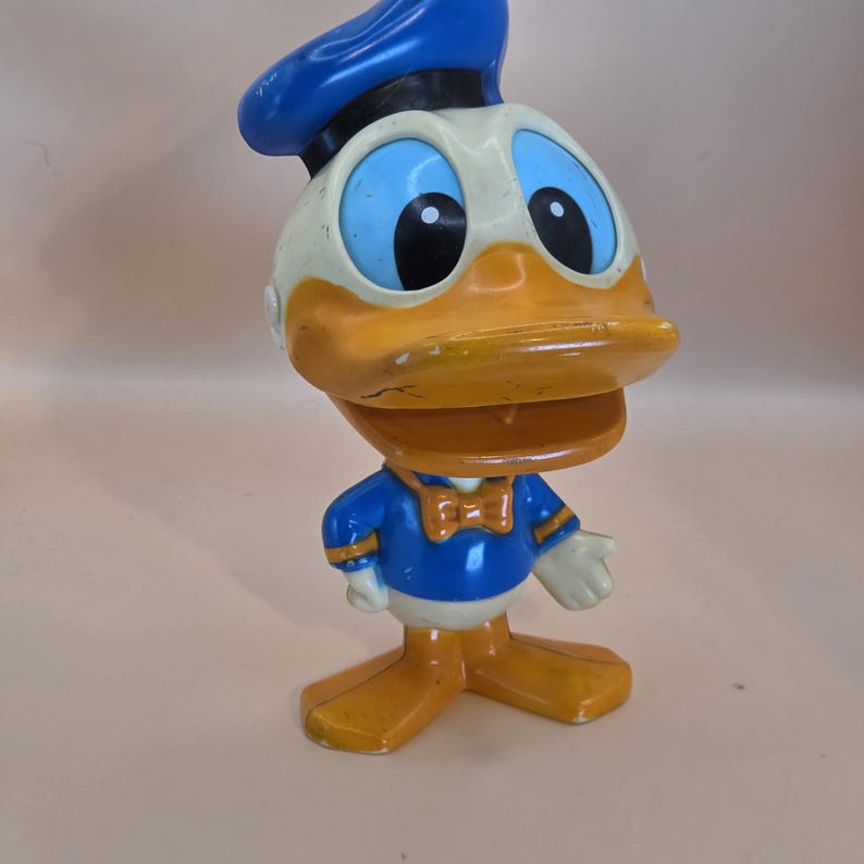 May include: A vintage Donald Duck figurine. The figure has a blue sailor hat, a white face, and a large orange beak. He wears a blue sailor shirt with a bow tie and white gloves. The figurine is standing on orange feet.