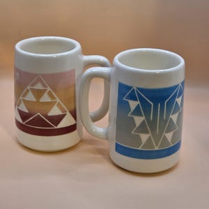 Peut inclure: Deux mugs en céramique blanche avec des anses. L'un des mugs présente un motif géométrique dans les tons de rose, d'orange et de marron. L'autre mug a un motif géométrique bleu et gris. Les deux mugs ont une forme cylindrique.