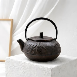 May include: A dark brown cast iron teapot with a black handle and lid. The teapot has a textured surface with a bamboo leaf design. The teapot is sitting on a white, textured surface.