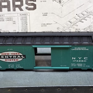 May include: A green model train boxcar with a black roof and ladder details. The side features the New York Central System logo, the text Cushion Underframe, and the identification number NYC 174280. The car has an open sliding door section.