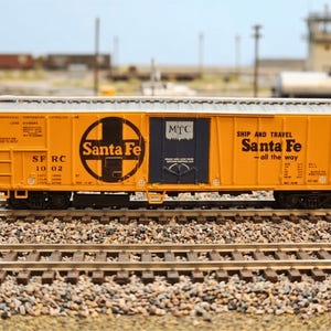 Puede incluir: Un vagón de mercancías Santa Fe amarillo con letras y logotipos negros. El vagón tiene el logotipo de Santa Fe y el texto "SHIP AND TRAVEL Santa Fe - all the way". El vagón de tren modelo está sobre una vía férrea en miniatura.