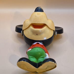 May include: Vintage Mickey Mouse toy figurine with a yellow face, black hat and body, and green pants. The toy has a red shirt and black shoes. The figurine is in a sitting position and has a smiling expression.