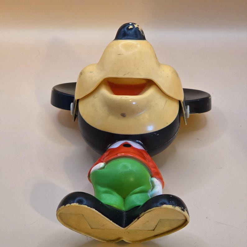 May include: Vintage Mickey Mouse toy figurine with a yellow face, black hat and body, and green pants. The toy has a red shirt and black shoes. The figurine is in a sitting position and has a smiling expression.