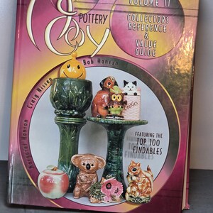 May include: A hardcover book titled McCoy Pottery Volume II Collectors Reference & Value Guide by Bob Hanson, Craig Nissen, and Margaret Hanson. The cover features a collection of ceramic figurines, including a yellow smiley face, animals, and an apple.