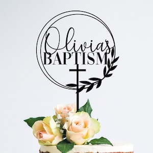 May include: Black cake topper with a circle design and the text "Olivia's Baptism" with a cross and leaves.