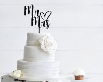 Op de afbeelding: Een drielaagse witte bruidstaart met een zwarte "Mr & Mrs"-topper en een witte roos op de bovenste laag. De taart staat op een witte taartstandaard en er zijn cupcakes op witte standaards op de achtergrond.