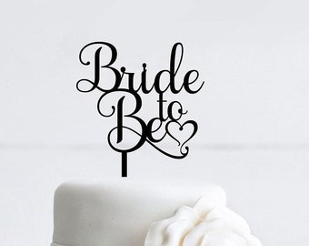 May include: A white two-tiered cake with a black acrylic cake topper that says "Bride to Be" with a heart. The cake is decorated with a white fondant rose.