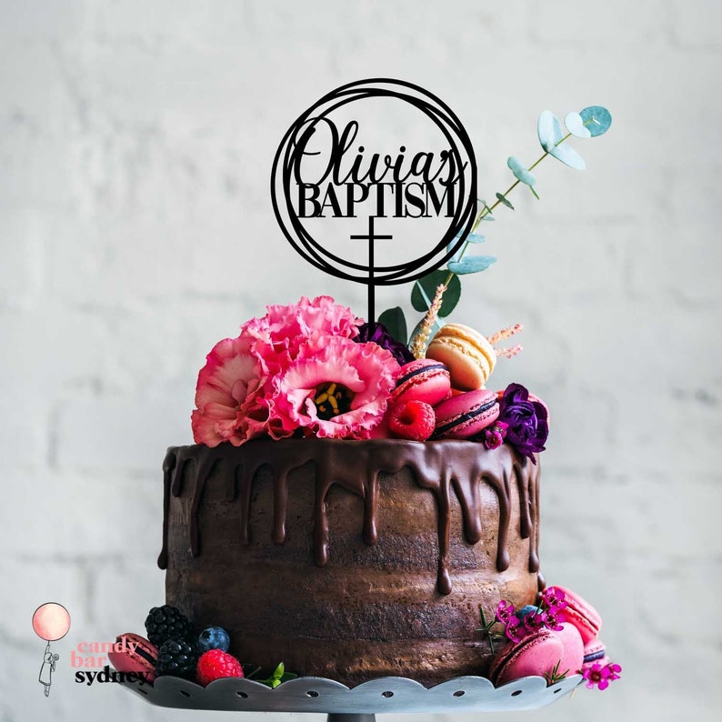 May include: Black acrylic cake topper with the text "Olivia's Baptism" in a circle with a cross. The topper is on a chocolate cake decorated with pink and purple flowers, macarons, and berries.