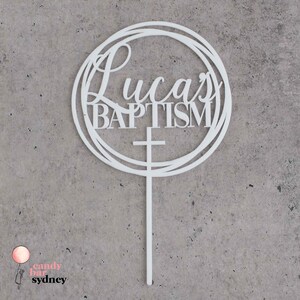 May include: White acrylic cake topper with a circle design and the text "Lucas Baptism" with a cross underneath.