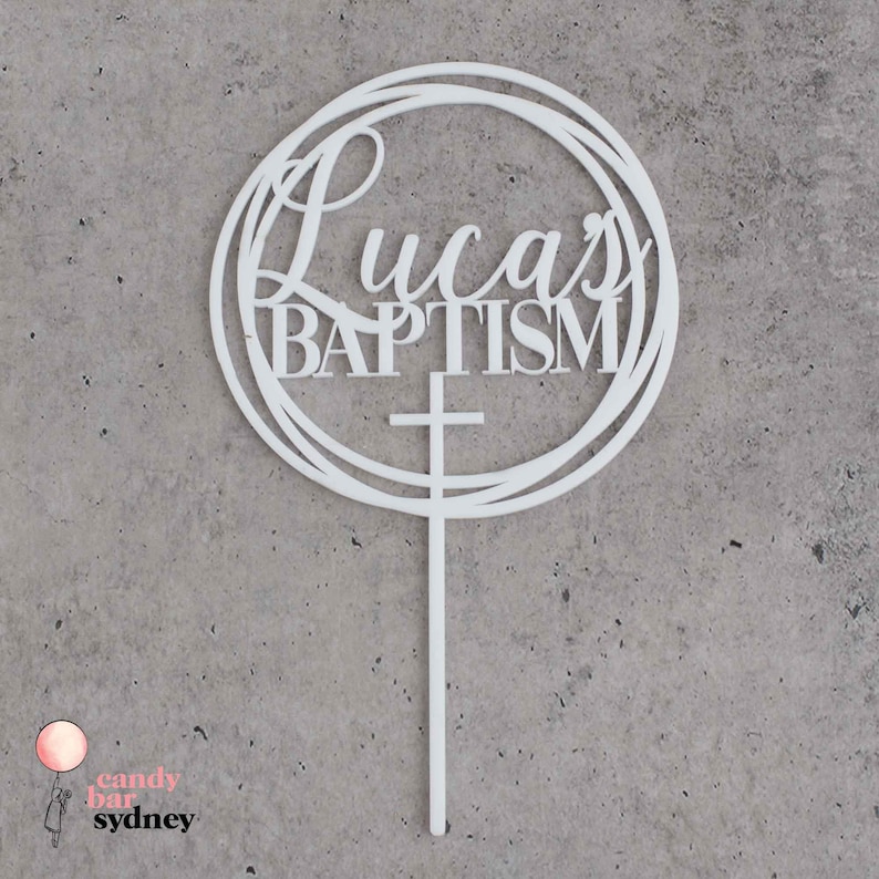 May include: White acrylic cake topper with a circle design and the text "Lucas Baptism" with a cross underneath.