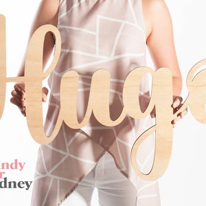 May include: A wooden sign with the word "hug" written in a cursive font. The sign is being held by a person wearing a light pink and white patterned top.