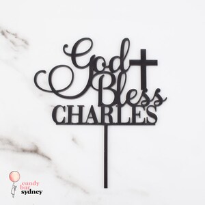 Op de afbeelding: Zwarte taarttopper met de tekst "God Bless Charles" en een kruisontwerp. De topper is gemaakt van zwart materiaal en is bedoeld om in een taart te steken. Een decoratief item voor een religieuze viering.