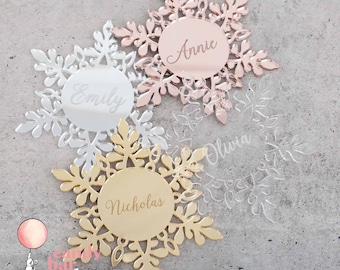 May include: Four snowflake ornaments with personalized names. The ornaments are made of mirrored acrylic in silver, gold, and rose gold. The names on the ornaments are Emily, Annie, Olivia, and Nicholas.