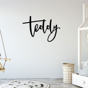 May include: A white wall with a black wooden sign that says "Teddy" in cursive script. There is a white cot with a white elephant head on the wall behind it. There is a wooden swing with two stuffed animals on it.
