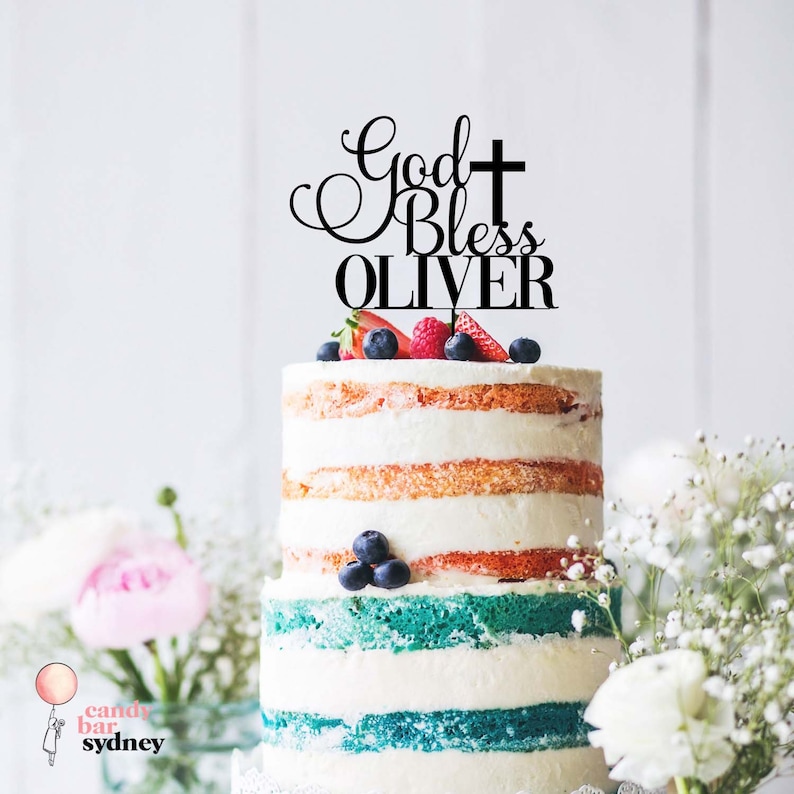 Op de afbeelding: Een meerlagige taart met witte frosting en kleurrijke lagen, gegarneerd met vers fruit en een zwarte taarttopper. De topper luidt "God Bless OLIVER" met een kruis.