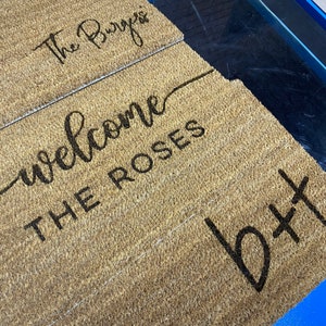 May include: A brown coir doormat with the text "The Burges" on the top half and "Welcome The Roses" on the bottom half. The text is in black and the doormat is on a blue surface.