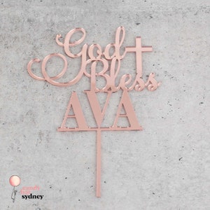 Op de afbeelding: Ros&eacute;gouden acryl taarttopper met de woorden "God Bless AVA" in een scriptlettertype en blokletters. De topper bevat een kruis en een steel om in een taart te steken. De achtergrond is een lichtgrijze ondergrond.