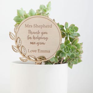 May include: A wooden plant stake with a circular sign that reads "Mrs. Shepherd Thank you for helping me grow Love Emma". The stake is decorated with a leafy design and is placed in a white pot with a succulent plant.