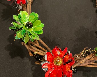 May include: A red and green floral wreath made from recycled materials. The wreath is decorated with small red and black beads.