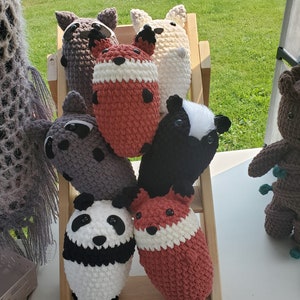 Pode incluir: Uma coleção de animais de pelúcia de crochê, incluindo um guaxinim, uma raposa, um panda, um gambá e um animal branco com olhos pretos. Os animais estão dispostos em uma prateleira de madeira.